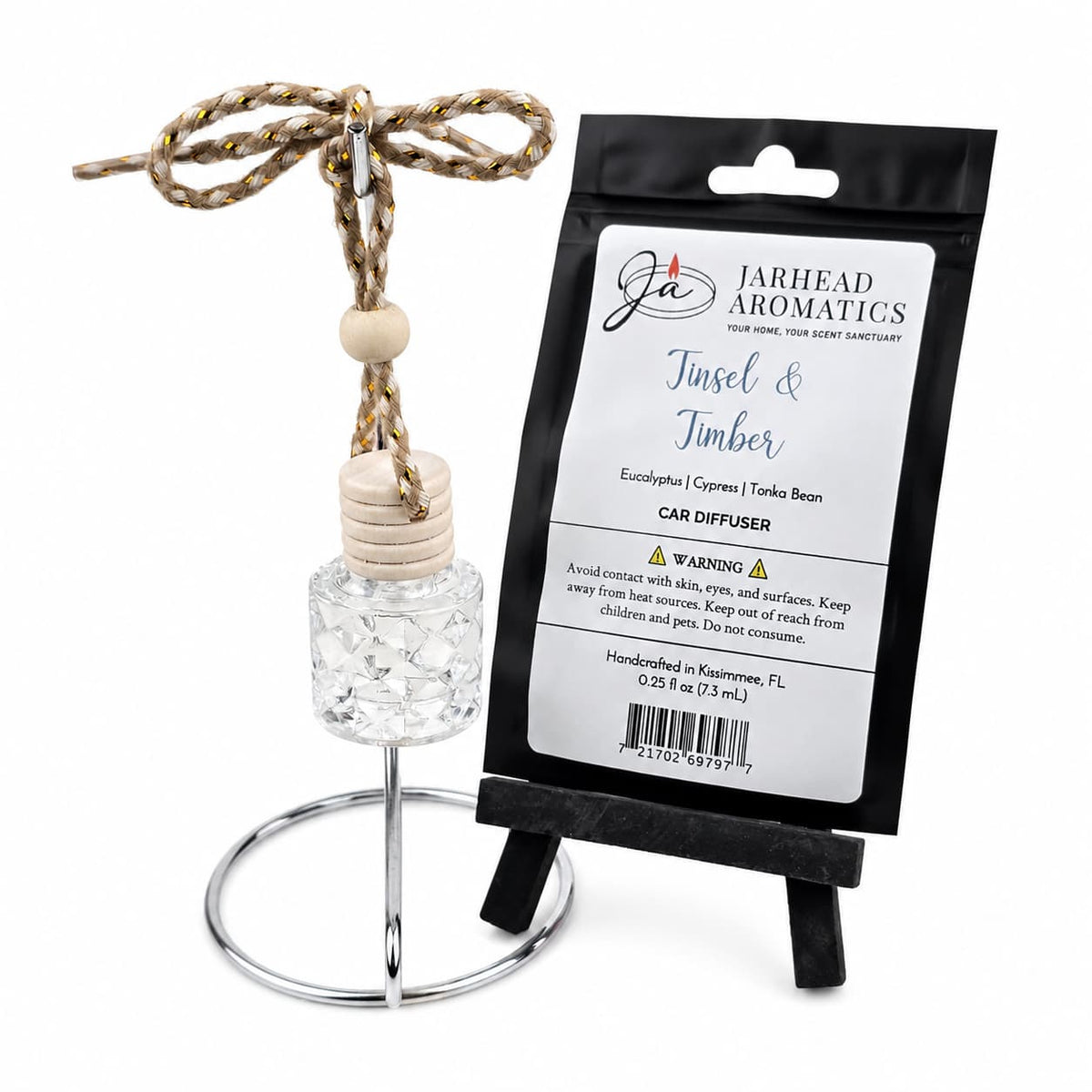 A glass Tinsel & Timber Car Diffuser with a wooden cap hangs from a metallic stand next to its black retail packaging.