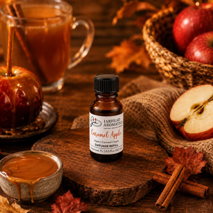 A bottle of Jarhead Aromatics Caramel Apple Cold Air Diffuser Refill | Crisp Apple, Rich Caramel & Warm Spice is surrounded by caramel, apples, a caramel apple, a spiced drink, cinnamon sticks, and autumn leaves on a rustic wooden table.