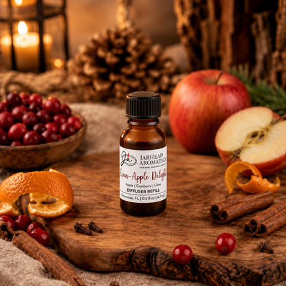 A bottle of Jarhead Aromatics Cran-Apple Delight Cold Air Diffuser Refill, featuring notes of cranberry, apple, and warm cinnamon, sits on a wooden board surrounded by apples, cranberries, orange peels, cinnamon sticks, and pinecones in a cozy, rustic setting with candles glowing in the background.