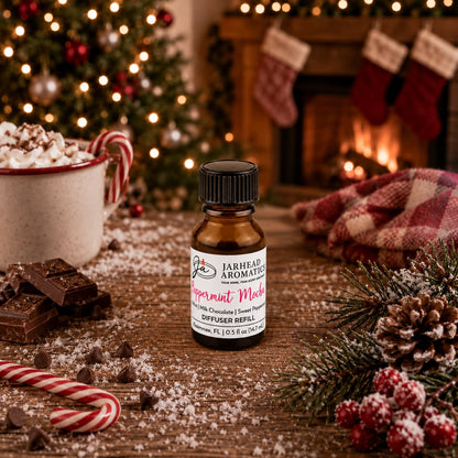 A brown glass bottle of Jarhead Aromatics Peppermint Mocha Cold Air Diffuser Refill sits on a wooden surface surrounded by chocolate, peppermint candies, and a mug of hot cocoa, with a Christmas tree and fireplace in the background.