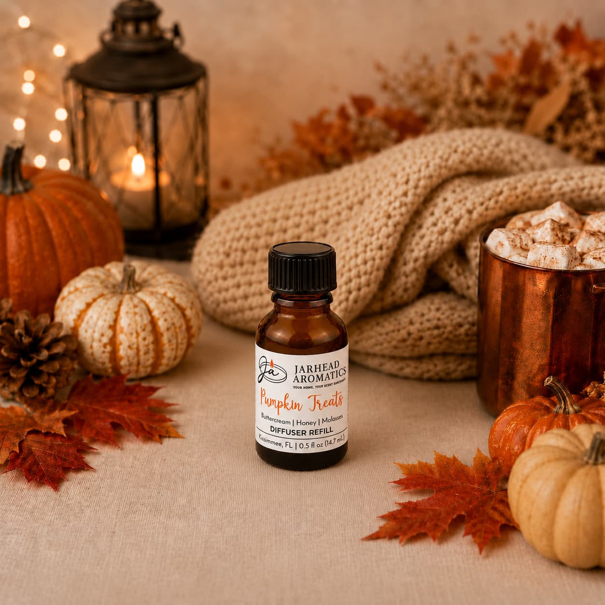 A small bottle of Jarhead Aromatics’ Pumpkin Treats Cold Air Diffuser Refill (Pumpkin, Caramel & Maple Pecan) rests on a table surrounded by autumn leaves, pumpkins, pinecones, a lantern, a copper mug topped with whipped cream, and a cozy beige knit blanket—perfect for filling your space with an inviting fall fragrance.