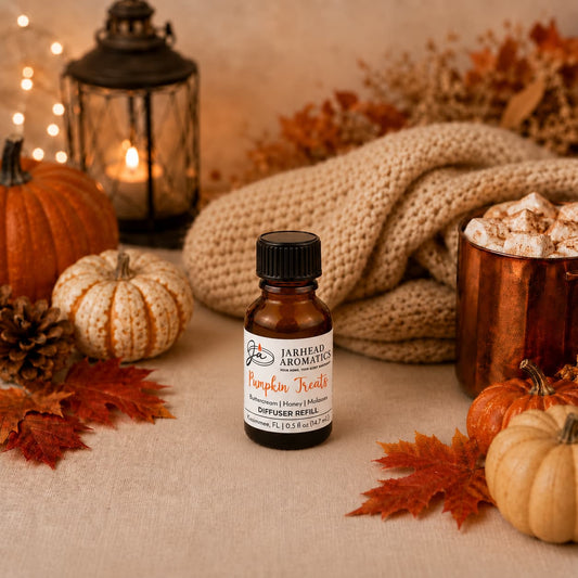A small bottle of Jarhead Aromatics’ Pumpkin Treats Cold Air Diffuser Refill (Pumpkin, Caramel & Maple Pecan) rests on a table surrounded by autumn leaves, pumpkins, pinecones, a lantern, a copper mug topped with whipped cream, and a cozy beige knit blanket—perfect for filling your space with an inviting fall fragrance.