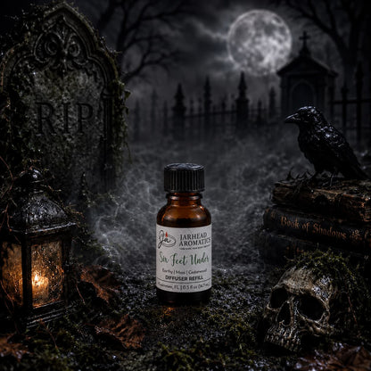 A small bottle of Jarhead Aromatics’ Six Feet Under Cold Air Diffuser Refill (featuring notes of damp earth, moss, and ancient oak) rests on mossy ground in a spooky graveyard—surrounded by a lantern, skull, raven, and tombstone—with fog and a full moon in the background; an eerie Halloween scent perfect for earthy home fragrance or as a cold air diffuser refill.