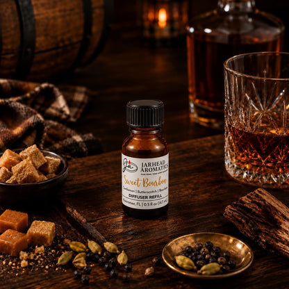 A bottle of Jarhead Aromatics Sweet Bourbon Cold Air Diffuser Refill, featuring notes of bourbon, butterscotch, and brown sugar, rests on a wooden table alongside a whiskey glass, brown sugar cubes, a decanter, spices, and a plaid cloth—creating a cozy atmosphere that perfectly embodies the inviting essence of this home fragrance oil.