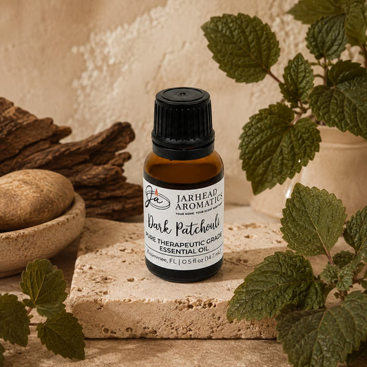 A small amber glass bottle of Jarhead Aromatics Dark Patchouli Essential Oil | Pure Pogostemon cablin Aromatherapy Oil rests on a stone surface, surrounded by green leaves, smooth stones, and textured wood. Celebrated for its therapeutic-grade purity and deep, earthy scent, this essential oil brings the essence of nature into your space.