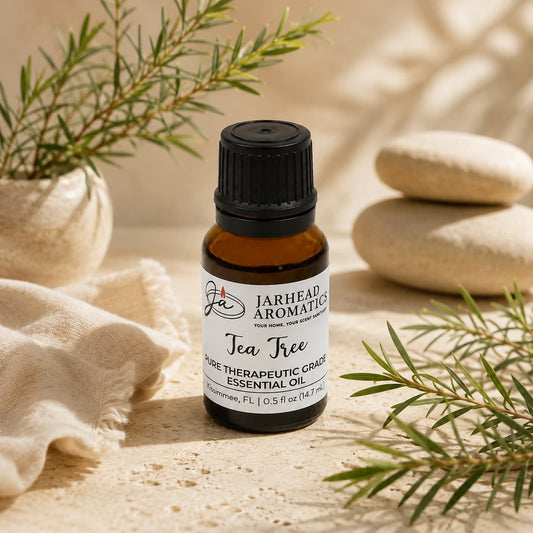 A small amber bottle of Jarhead Aromatics Tea Tree Essential Oil | Pure Melaleuca alternifolia Aromatherapy Oil is displayed on a beige surface, surrounded by green sprigs, smooth white stones, and a folded cream cloth, creating an inviting aromatherapy setting.