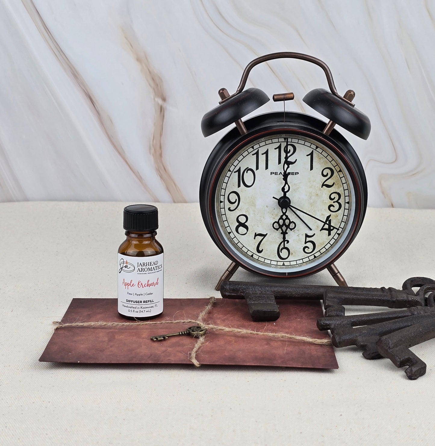 A vintage alarm clock, a brown string-tied envelope, a Jarhead Aromatics Apple Orchard - Cold Air Diffuser Refill bottle, and three antique keys are arranged on a light surface against a marble background.