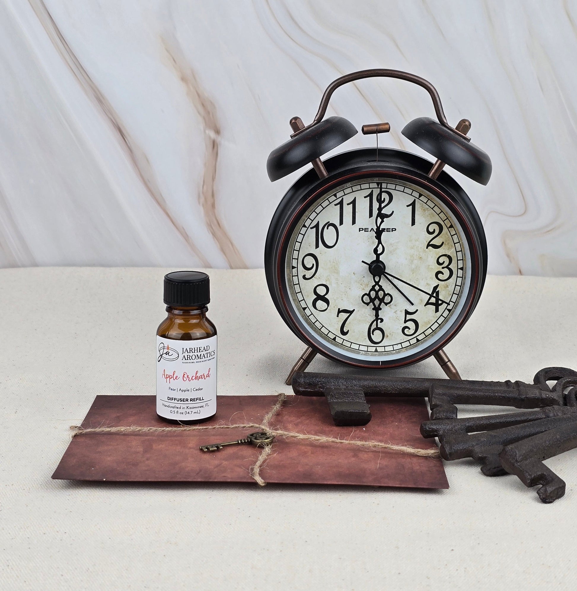 A vintage alarm clock, a brown string-tied envelope, a Jarhead Aromatics Apple Orchard - Cold Air Diffuser Refill bottle, and three antique keys are arranged on a light surface against a marble background.