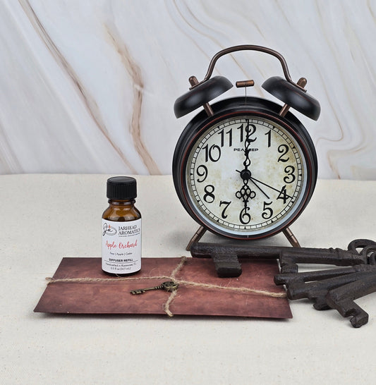 A vintage alarm clock, a brown string-tied envelope, a Jarhead Aromatics Apple Orchard - Cold Air Diffuser Refill bottle, and three antique keys are arranged on a light surface against a marble background.