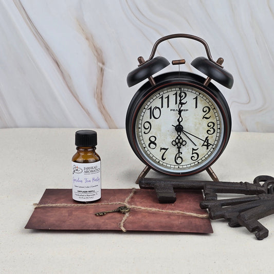 A Garden Tea Party Cold Air Diffuser Refill by Jarhead Aromatics, a vintage-style alarm clock, two large metal keys, and a tied brown envelope are displayed on a light surface with a marble background.