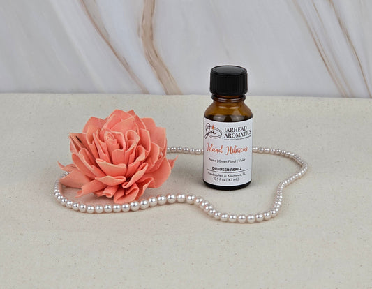 A small brown bottle labeled Island Hibiscus - Cold Air Diffuser Refill by Jarhead Aromatics sits on a light surface beside a coral artificial flower and a looped pearl necklace, with a marble-patterned background behind them.