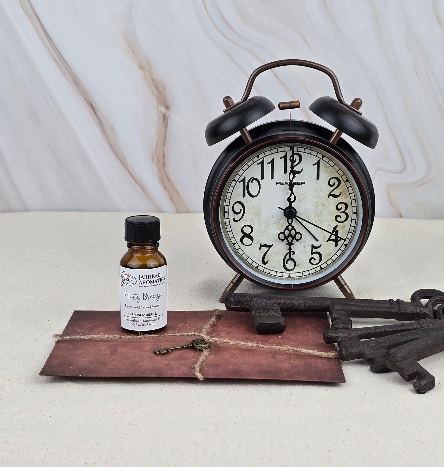 A vintage black alarm clock displays 12:02 on a beige surface beside Jarhead Aromatics Minty Breeze - Cold Air Diffuser Refill, an old envelope tied with twine, and two large antique metal keys. The backdrop is light marble.