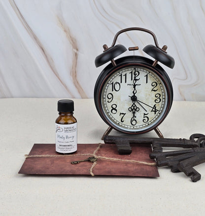 A vintage black alarm clock displays 12:02 on a beige surface beside Jarhead Aromatics Minty Breeze - Cold Air Diffuser Refill, an old envelope tied with twine, and two large antique metal keys. The backdrop is light marble.