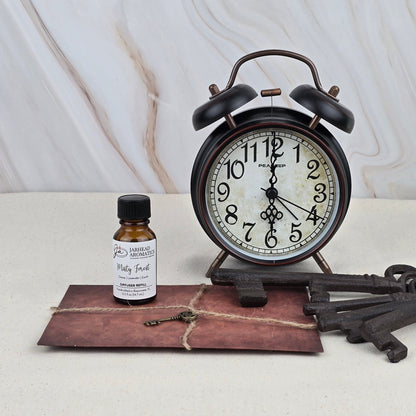 A vintage alarm clock stands beside a small bottle of Jarhead Aromatics Misty Forest - Cold Air Diffuser Refill, an old brown envelope tied with twine, and two antique keys, all arranged on a light surface against a marble background.