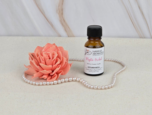 A small amber bottle labeled “Mystic Orchid - Cold Air Diffuser Refill” by Jarhead Aromatics rests near a pink artificial flower and pearls, evoking the exotic orchid scent on a cream surface with a marbled white and beige background.