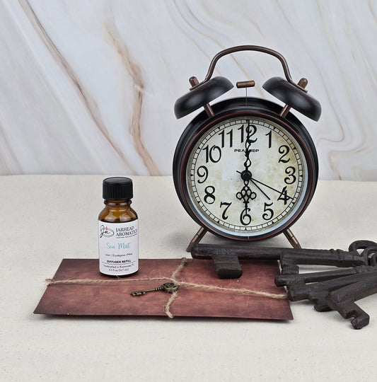 A Jarhead Aromatics Sea Mist Cold Air Diffuser Refill sits beside a vintage black alarm clock, an old key, and a tied brown envelope on a light surface with a marble-patterned background.