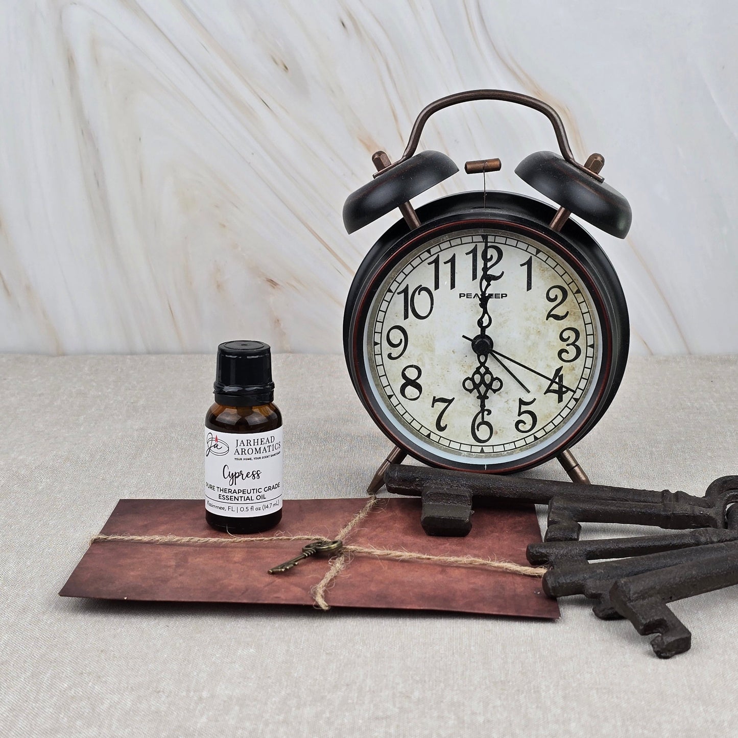 A vintage black alarm clock, three antique keys, a brown twine-tied envelope, and a bottle of Jarhead Aromatics Cypress Pure Therapeutic-Grade Essential Oil are arranged on a textured surface with a marble-patterned background.