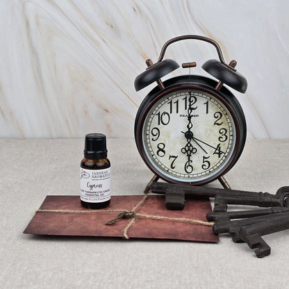 A vintage black alarm clock, three antique keys, a brown twine-tied envelope, and a bottle of Jarhead Aromatics Cypress Pure Therapeutic-Grade Essential Oil are arranged on a textured surface with a marble-patterned background.