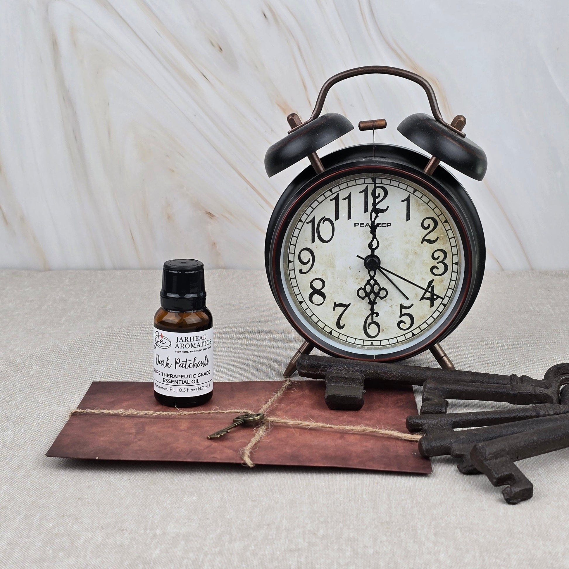A vintage alarm clock sits beside a bottle of Jarhead Aromatics Dark Patchouli Pure Therapeutic-Grade Essential Oil, a tied brown envelope, and three antique keys on beige with a marble-patterned backdrop, evoking calming aromatherapy.