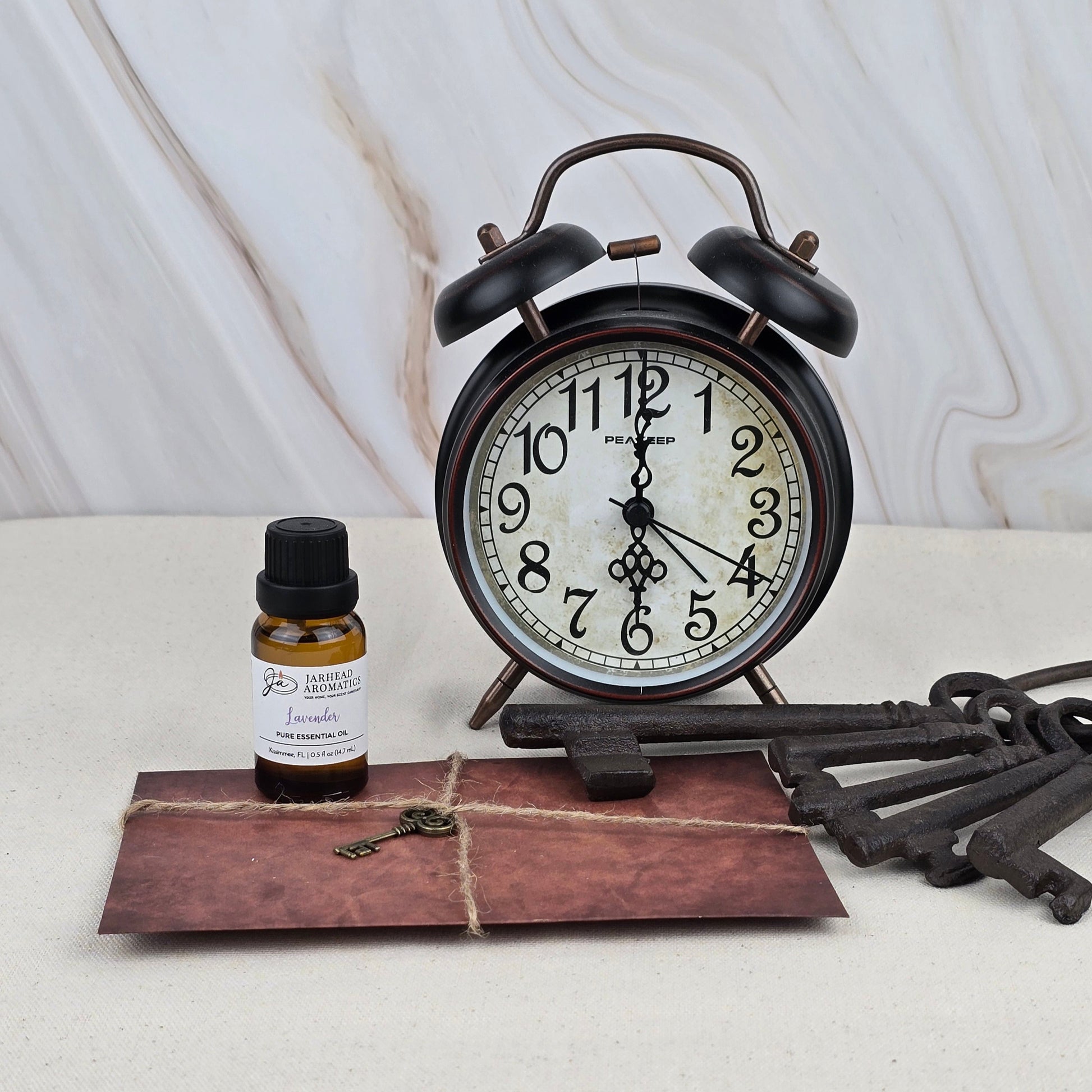 A black vintage alarm clock, Jarhead Aromatics Lavender - Pure Essential Oil, a tied brown envelope with a key, and three large antique keys sit on a light surface against marble—ideal for aromatherapy lovers.