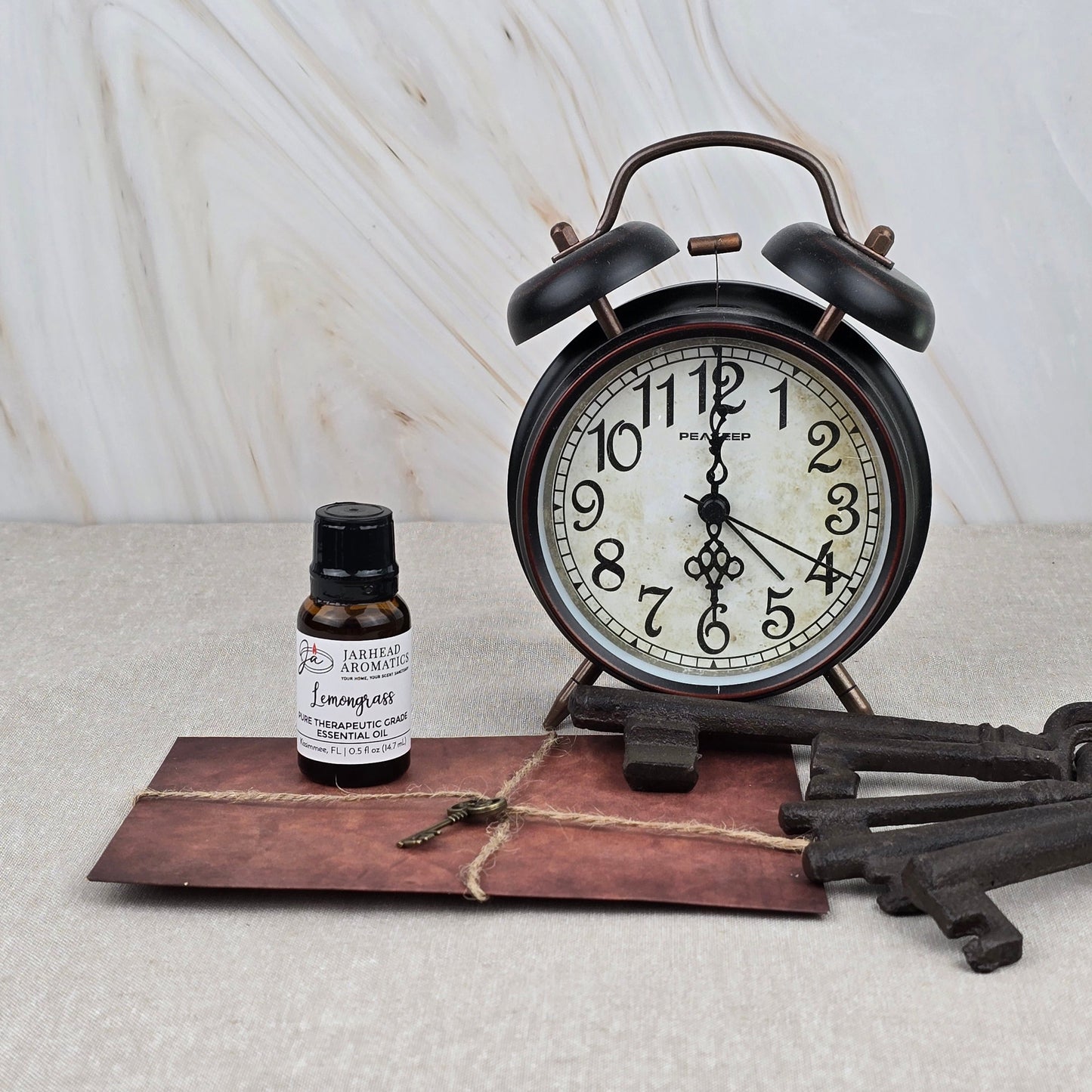 A vintage alarm clock, Jarhead Aromatics’ Lemongrass Pure Therapeutic-Grade Essential Oil, an old brown envelope, and three antique keys sit on a light surface against a marble-like background.