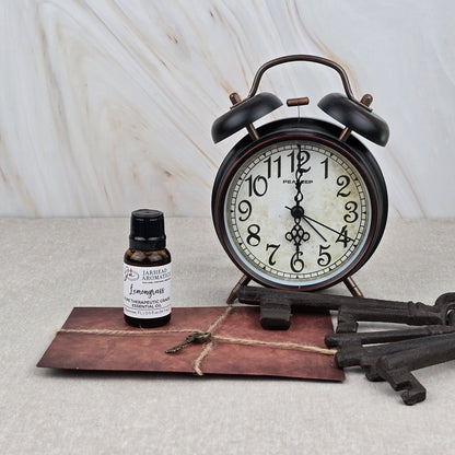 A vintage alarm clock, Jarhead Aromatics’ Lemongrass Pure Therapeutic-Grade Essential Oil, an old brown envelope, and three antique keys sit on a light surface against a marble-like background.