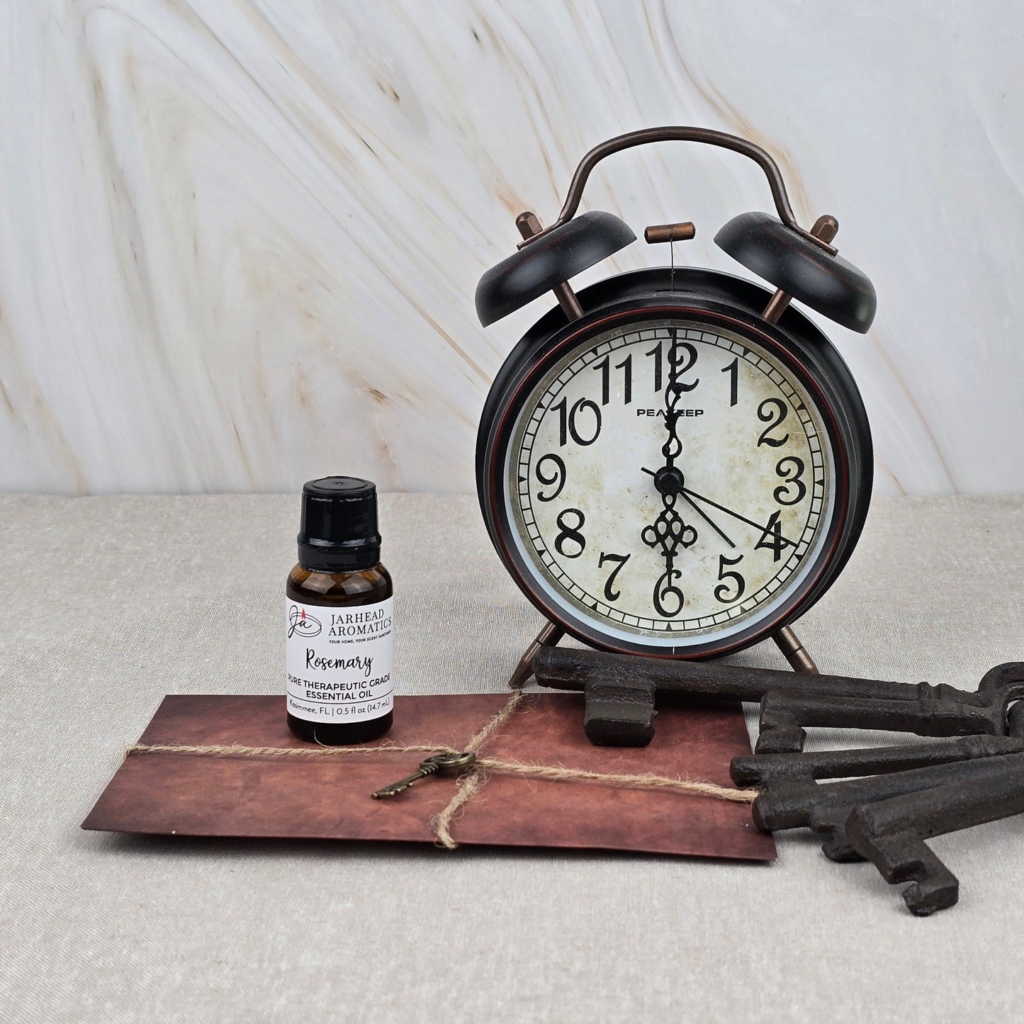 A vintage alarm clock, a brown envelope with string, Rosemary Pure Therapeutic-Grade Essential Oil by Jarhead Aromatics, and two antique-style keys rest on beige against a marble-patterned background.