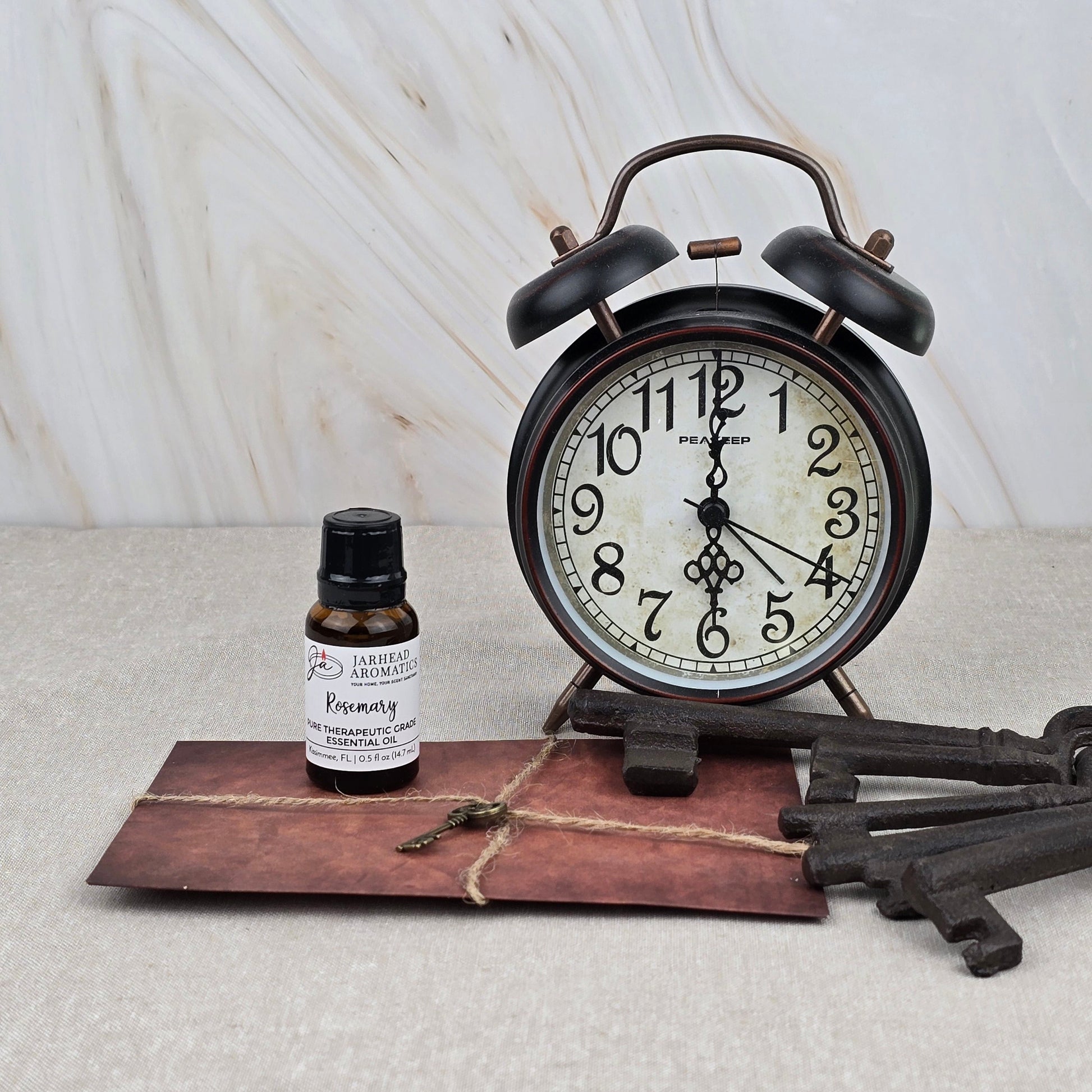 A vintage alarm clock, a brown envelope with string, Rosemary Pure Therapeutic-Grade Essential Oil by Jarhead Aromatics, and two antique-style keys rest on beige against a marble-patterned background.