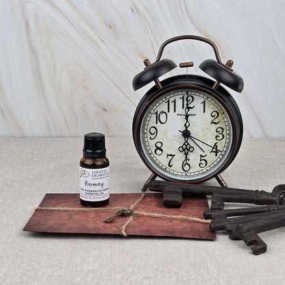 A vintage alarm clock, a brown envelope with string, Rosemary Pure Therapeutic-Grade Essential Oil by Jarhead Aromatics, and two antique-style keys rest on beige against a marble-patterned background.