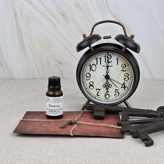 A vintage alarm clock, a brown envelope with string, Rosemary Pure Therapeutic-Grade Essential Oil by Jarhead Aromatics, and two antique-style keys rest on beige against a marble-patterned background.