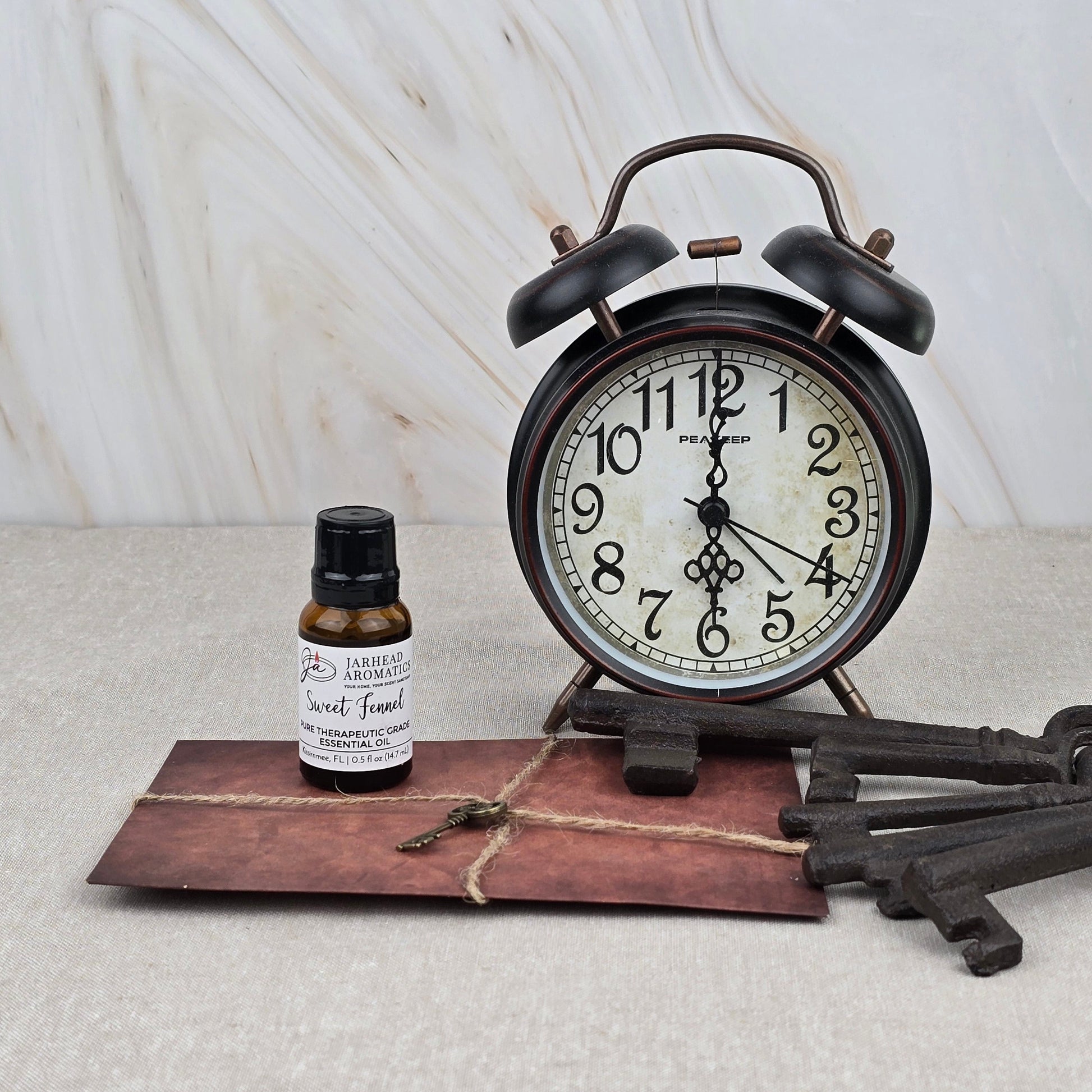 A vintage alarm clock shows 10:12 beside Jarhead Aromatics’ Sweet Fennel Pure Therapeutic-Grade Essential Oil, a twine-tied brown envelope, and two antique keys on beige with a marble-patterned background.