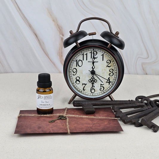 A vintage alarm clock, three large metal keys, and a brown envelope tied with string sit beside a small bottle of Jarhead Aromatics Tea Tree - Pure Essential Oil on a light surface against a marble-patterned background—ideal for aromatherapy.
