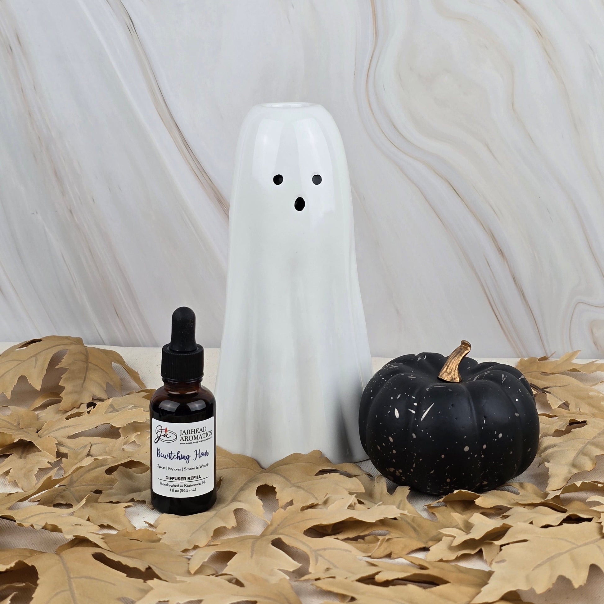 A refill of Bewitching Hour car diffuser by Jarhead Aromatics sits with a white ceramic ghost and a black pumpkin with gold stem on tan leaves, all set against a marble-patterned background for Halloween.