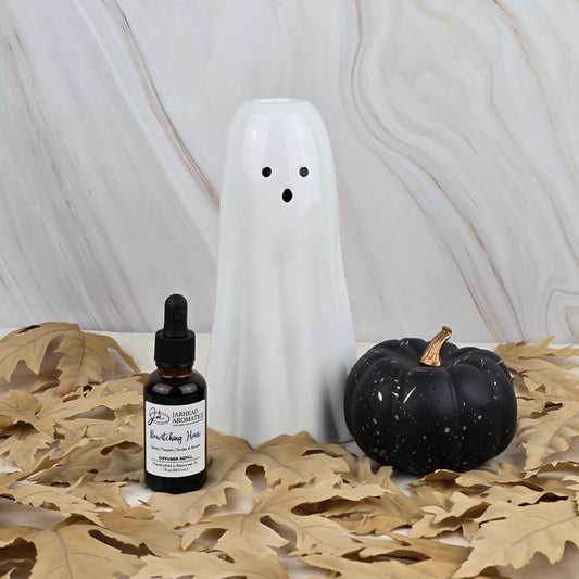 A refill of Bewitching Hour car diffuser by Jarhead Aromatics sits with a white ceramic ghost and a black pumpkin with gold stem on tan leaves, all set against a marble-patterned background for Halloween.
