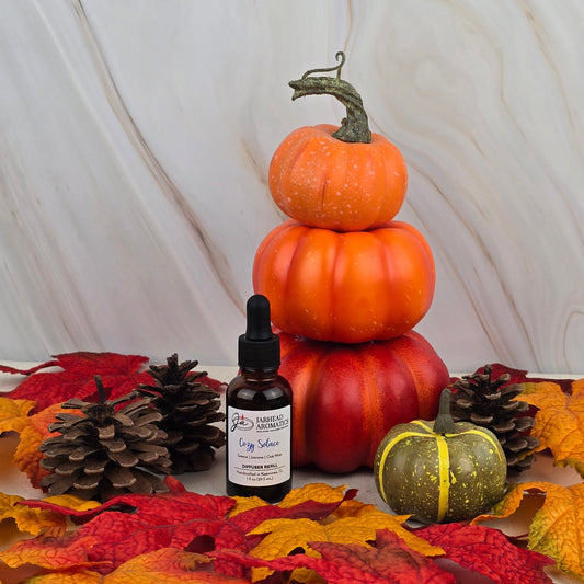 Three stacked orange pumpkins, a small green pumpkin, pinecones, and the Cozy Solace Car Diffuser Refill by Jarhead Aromatics are surrounded by autumn leaves, evoking oak moss on a light marble background.