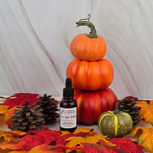 A stack of three small pumpkins, a green pumpkin, pinecones, and autumn leaves surround a bottle of Jarhead Aromatics Cran-Apple Delight Car Diffuser Refill from the Fall Collection, all set against a marble background.