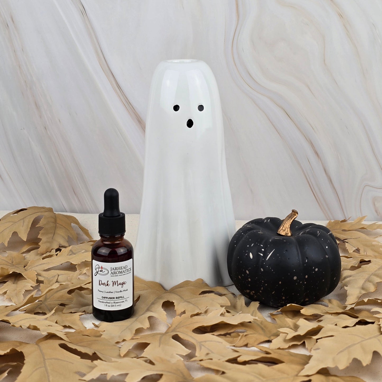 A ceramic ghost, a black pumpkin, and a bottle of Jarhead Aromatics Dark Magic Car Diffuser Refill from the Halloween Collection rest on dried tan leaves atop a light marble background.