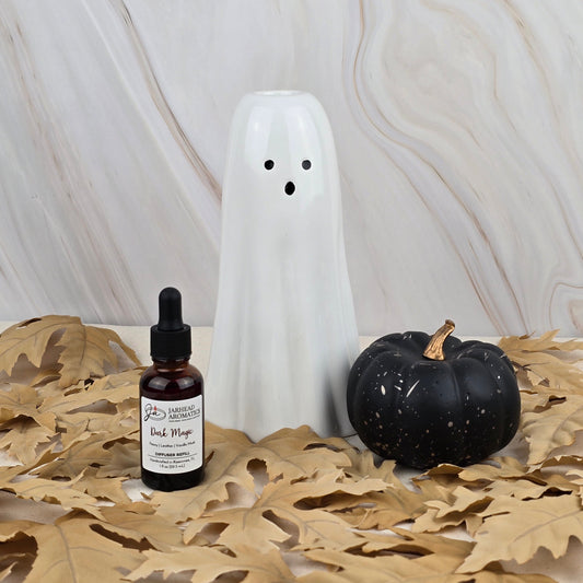 A ceramic ghost, a black pumpkin, and a bottle of Jarhead Aromatics Dark Magic Car Diffuser Refill from the Halloween Collection rest on dried tan leaves atop a light marble background.