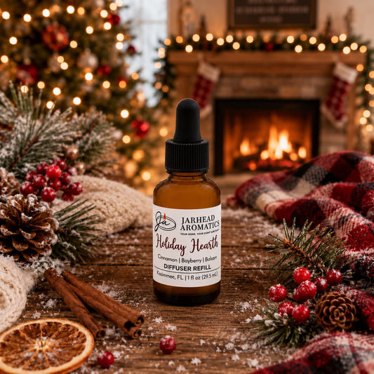 Amber glass Holiday Hearth Car Diffuser Refill bottle from Jarhead Aromatics on a wooden surface with cinnamon sticks, dried citrus, pine branches, and berries, with a fireplace and Christmas tree in the background.