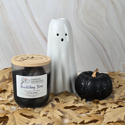 A white ceramic ghost, a black decorative pumpkin, and Jarhead Aromatics’ Bewitching Hour Soy Wax Candle from the Halloween Collection, with a smoky woods scent, rest on brown leaves against a marbled backdrop.