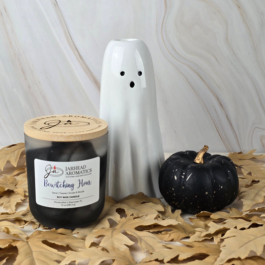 A white ceramic ghost, a black decorative pumpkin, and Jarhead Aromatics’ Bewitching Hour Soy Wax Candle from the Halloween Collection, with a smoky woods scent, rest on brown leaves against a marbled backdrop.