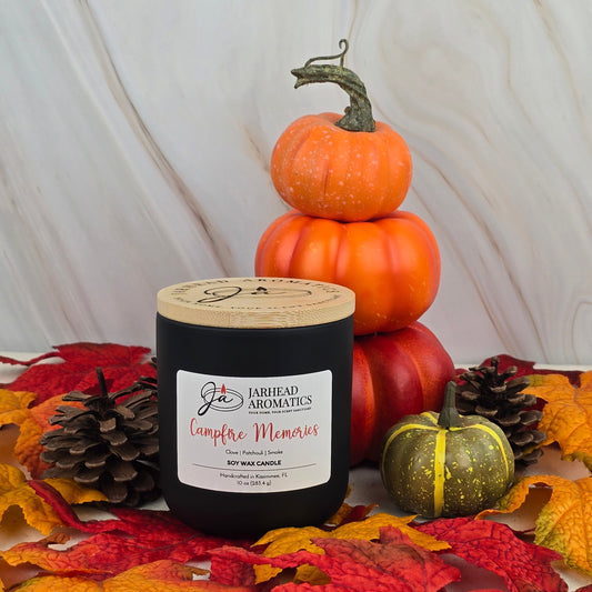 A black Jarhead Aromatics Campfire Memories - Soy Wax Candle - Fall Collection sits among autumn leaves, pine cones, mini pumpkins, and a green pumpkin, for a cozy fall scene with campfire fragrance and essential oils.