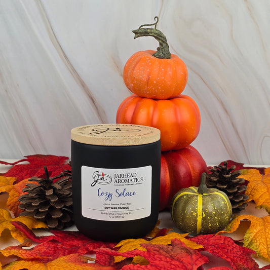 The Cozy Solace soy wax candle from Jarhead Aromatics’ Fall Collection rests on autumn leaves, pinecones, and pumpkins, filling the air with soothing jasmine, fresh greens, and oak moss scents against a light wood backdrop.