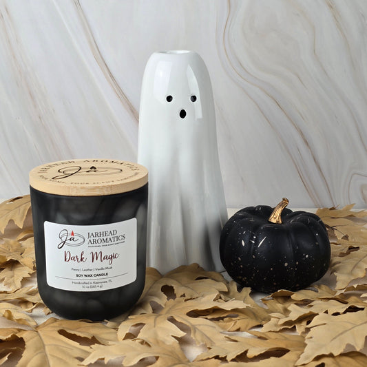 A Jarhead Aromatics Dark Magic - Soy Wax Candle with vanilla musk notes, a ceramic ghost, and a black pumpkin rest on dried leaves against a marble-patterned backdrop.