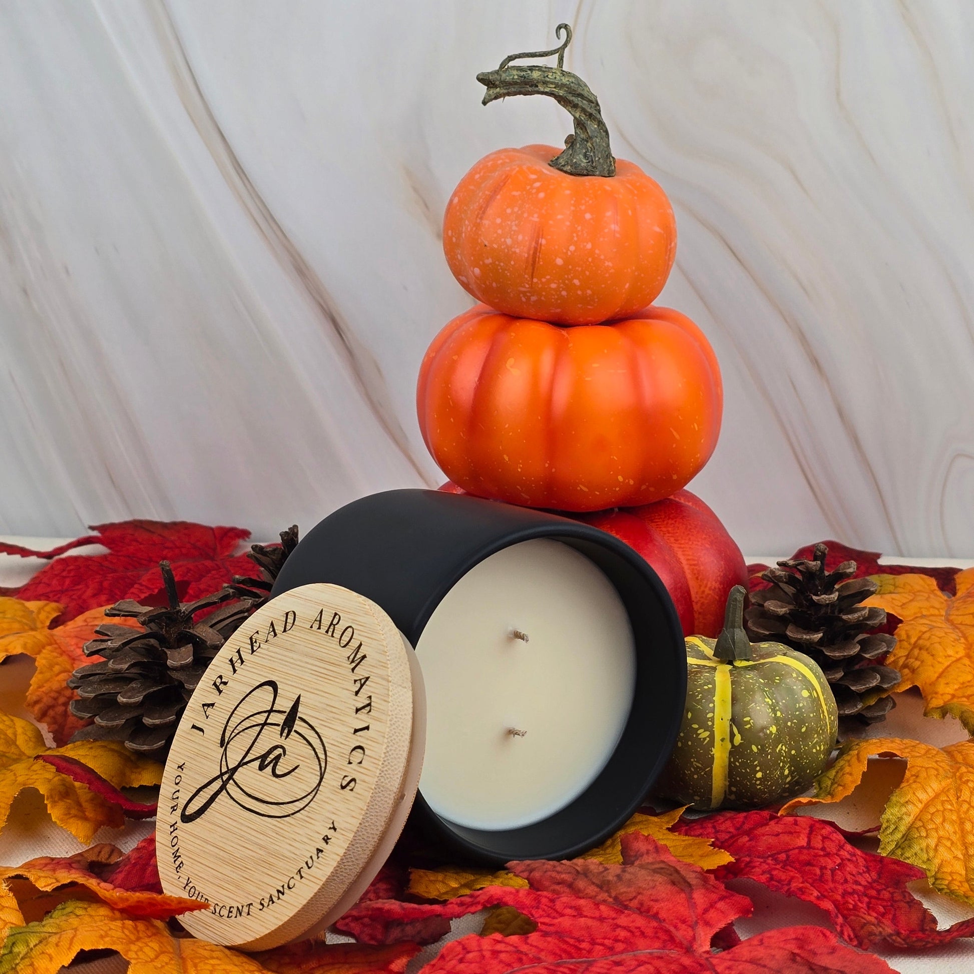 The Cozy Solace soy wax candle by Jarhead Aromatics, in a black jar with wooden lid, sits open among autumn leaves, pinecones, and small pumpkins. Oak moss and fresh greens blend as three stacked pumpkins create a fall scene in the background.