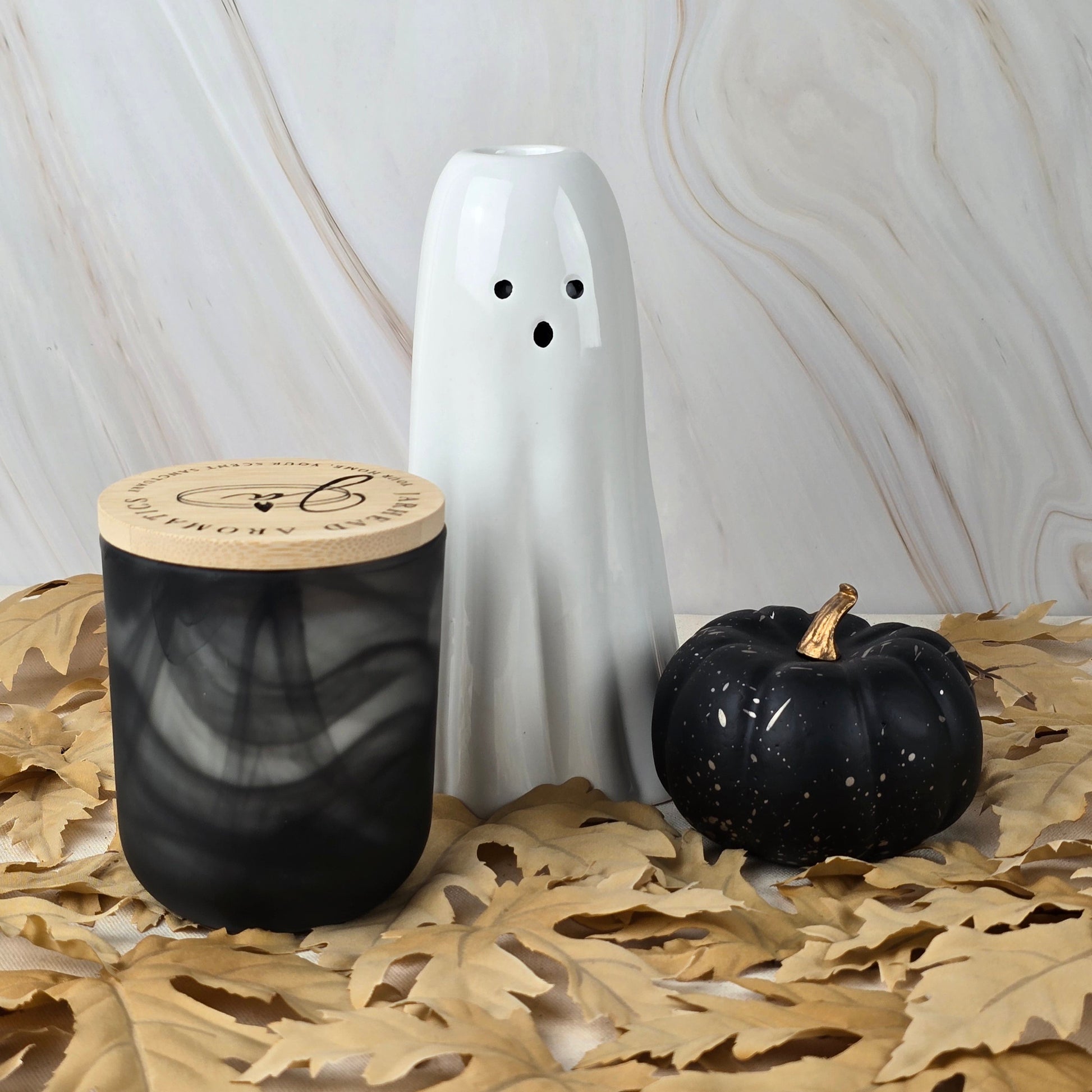 A white ceramic ghost, Jarhead Aromatics’ Six Feet Under soy wax candle—a black jar with a wooden lid and earthy moss scent—and a small black pumpkin sit on dried leaves against a marbled background.