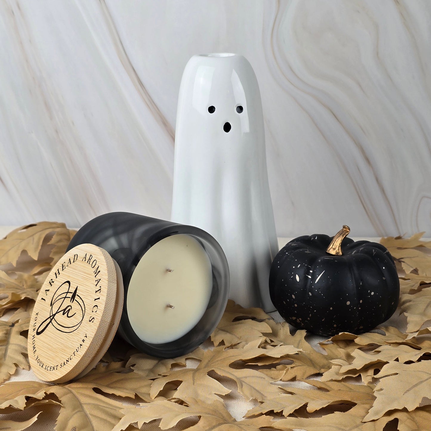 The Six Feet Under Soy Wax Candle by Jarhead Aromatics rests with a ghost ceramic and black pumpkin ornament on dried leaves against marble, capturing the mysterious aroma of an overgrown graveyard.