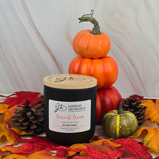 A black Jarhead Aromatics Harvest Cream Soy Wax Candle from the Fall Collection rests on autumn leaves with pinecones, a green pumpkin, and mini orange pumpkins in front of light marble, filling the air with a cozy fall scent.