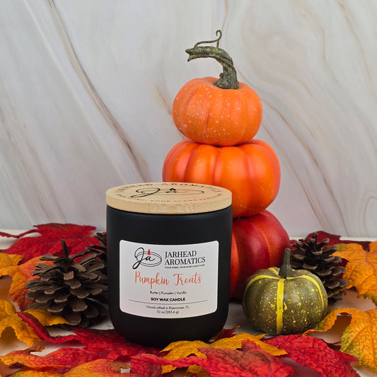 A black Pumpkin Treats - Soy Wax Candle from Jarhead Aromatics Fall Collection sits among autumn leaves, pine cones, and pumpkins, with a pumpkin stack in the background, capturing a cozy autumn vibe against a marble backdrop.