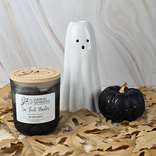 A Jarhead Aromatics Six Feet Under Soy Wax Candle, black and earthy, sits with a white ghost-shaped ceramic ornament and a small black pumpkin on dried leaves against a light marbled background.